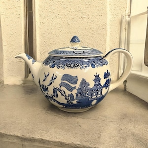 May include: A white ceramic teapot with a blue Willow pattern design. The teapot features a curved spout, a rounded body, and a matching lid with a small knob. The design includes scenes of pagodas, trees, and birds.