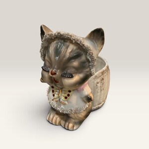 May include: A ceramic cat figurine planter featuring a grey and white kitten wearing a bonnet with glittery trim. The kitten has painted eyelashes, a small bow at the neck, and a textured basket-like container attached to its back for holding items.