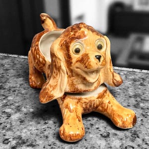 May include: A ceramic dog figurine with large, expressive eyes and floppy ears. The dog is tan and brown with a glossy finish. The figurine has a hollowed-out area on its back, suggesting it may be a planter or container.