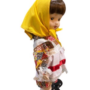 May include: A vintage porcelain doll with brown hair and a yellow headscarf. The doll wears a patchwork patterned dress with white lace trim and a white apron with red ribbon. Black shoes complete the outfit.
