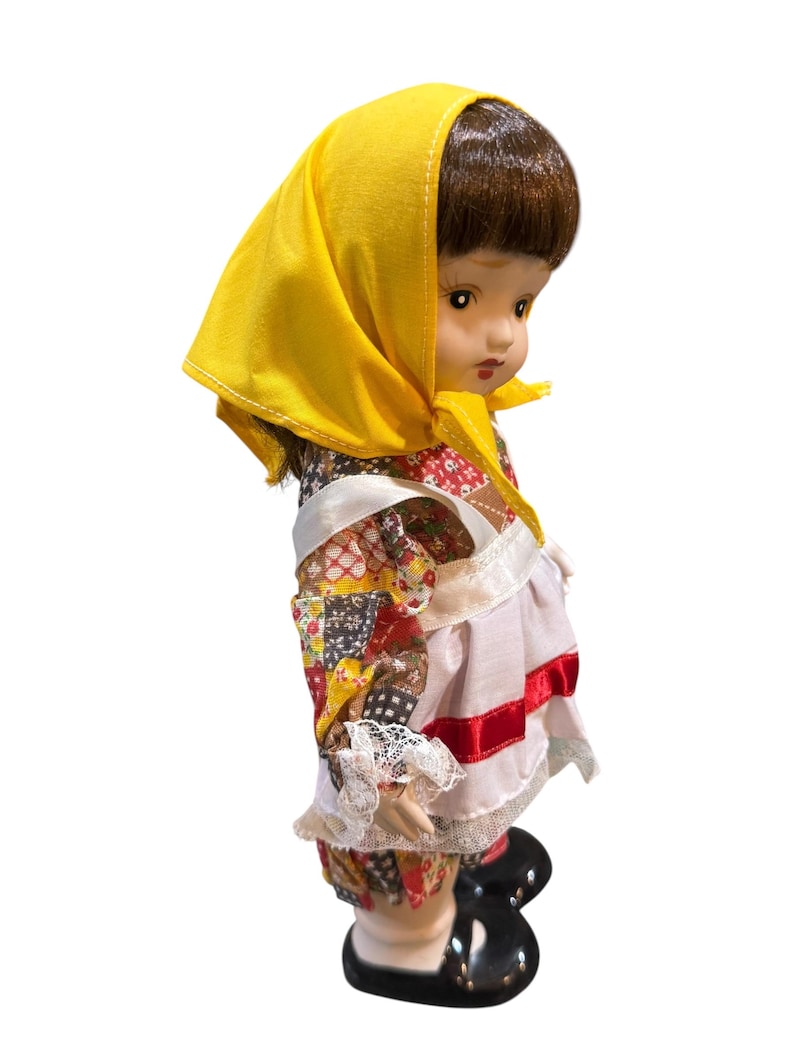 May include: A vintage porcelain doll with brown hair and a yellow headscarf. The doll wears a patchwork patterned dress with white lace trim and a white apron with red ribbon. Black shoes complete the outfit.