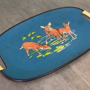 May include: Oval serving tray with a teal background and a black border. The tray features a painted scene of three deer in shades of brown and white, with green foliage and water lilies. The handles are wrapped in yellow woven material.
