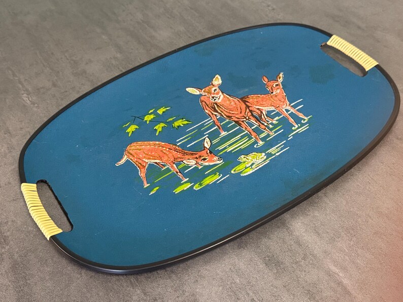 May include: Oval serving tray with a teal background and a black border. The tray features a painted scene of three deer in shades of brown and white, with green foliage and water lilies. The handles are wrapped in yellow woven material.