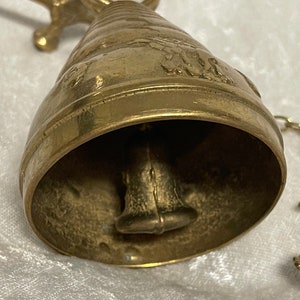 May include: A brass bell with a decorative design and a hanging chain. The bell has a small clapper inside and is attached to a metal stand.