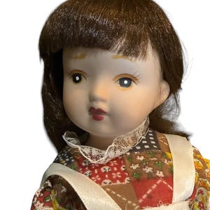 May include: A porcelain doll with brown hair and blue eyes. The doll wears a dress with a patchwork pattern in red, brown, and yellow, and a white apron. The doll has a delicate, painted face with red lips.