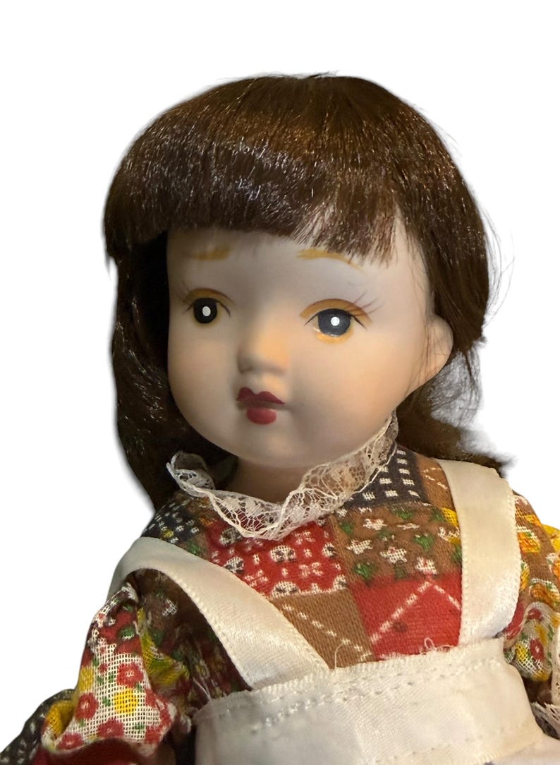 May include: A porcelain doll with brown hair and blue eyes. The doll wears a dress with a patchwork pattern in red, brown, and yellow, and a white apron. The doll has a delicate, painted face with red lips.
