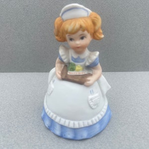 May include: A porcelain figurine of a girl wearing a white dress with blue trim and a white nurse's cap. She is holding a tray with a small cake on it.
