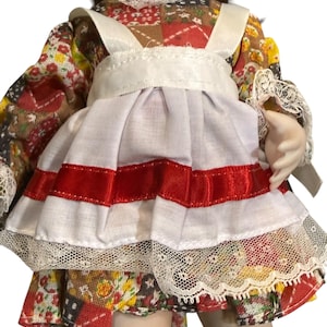 May include: A porcelain doll with brown hair and red lips. The doll wears a patchwork print dress with a white apron featuring red velvet stripes and lace trim. The doll's sleeves are also trimmed with lace.
