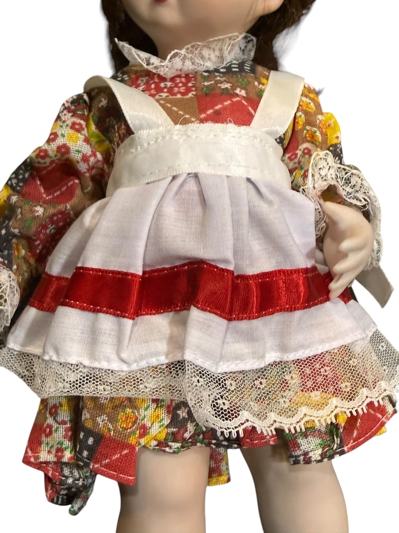 May include: A porcelain doll with brown hair and red lips. The doll wears a patchwork print dress with a white apron featuring red velvet stripes and lace trim. The doll's sleeves are also trimmed with lace.
