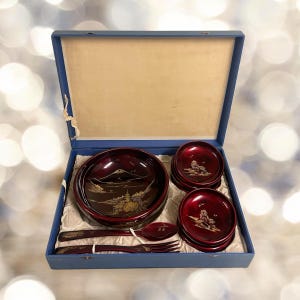 May include: A red lacquered Japanese bento box set with a mountain scene design. The set includes a large bowl, five smaller bowls, a spoon, and chopsticks. The box is lined with white fabric and has a blue exterior.