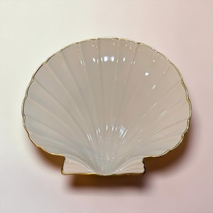 May include: A white ceramic dish shaped like a scallop shell, featuring a glossy finish and a thin, metallic gold trim along the scalloped edges. The bowl has deep, radiating ridges that mimic the natural texture of a seashell.