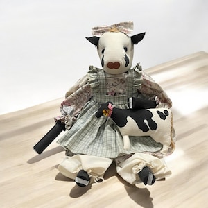 May include: A handmade cow doll wearing a green and white plaid dress with a floral print top and a pink bow. The doll is holding a smaller black and white cow doll.