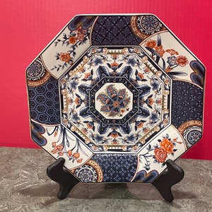 May include: Octagonal ceramic plate with a white background and intricate blue, gold, and orange floral patterns. The plate is on a black stand.