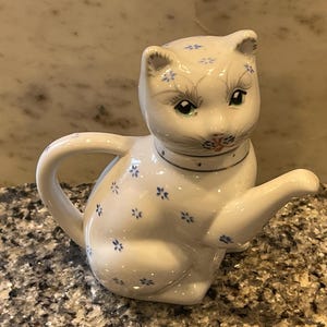 May include: White ceramic teapot shaped like a cat with blue and gold floral details. The teapot has a spout and a handle.