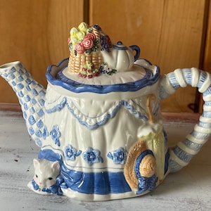 May include: A white ceramic teapot with a blue and white checkered pattern. The teapot is decorated with a basket of flowers, a small teapot, a straw hat, an umbrella, and a white cat.