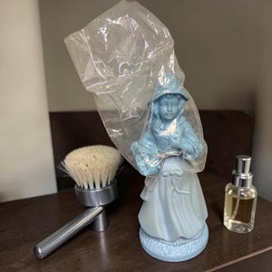 May include: A light blue figurine of a woman in a dress and hat, wrapped in plastic. A brush with a silver handle and a small perfume bottle are also visible. The figurine is the focal point, suggesting a collectible or decorative item.