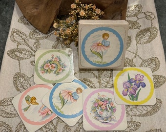 May include: Set of six vintage paper coasters with a pastel color scheme. Each coaster features a different illustration of a child, flowers, and a swan. The coasters are in a square box with a blue and white design.