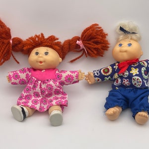 May include: Two Cabbage Patch Kids dolls, one with red yarn hair and a pink floral dress, the other with blonde yarn hair and a blue and white patterned shirt and blue pants.