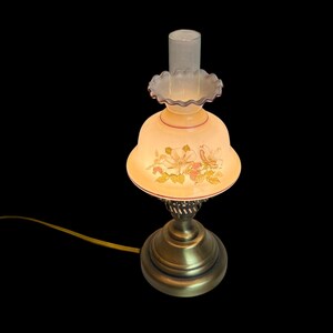 May include: A vintage-style table lamp with a floral design on the shade. The lamp has a brass base and a frosted glass shade with a ruffled edge. The shade features a painted floral pattern in shades of pink, yellow, and green.