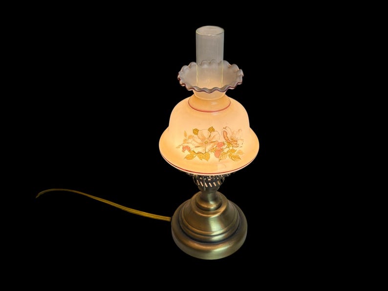 May include: A vintage-style table lamp with a floral design on the shade. The lamp has a brass base and a frosted glass shade with a ruffled edge. The shade features a painted floral pattern in shades of pink, yellow, and green.