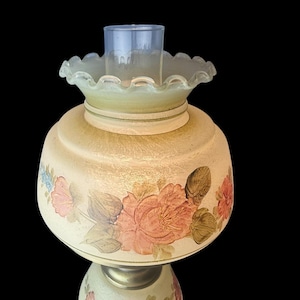 Puede incluir: Una l&aacute;mpara de aceite de estilo vintage con una base redondeada de color crema adornada con dise&ntilde;os florales rosas y melocot&oacute;n y hojas verdes. La l&aacute;mpara presenta una pantalla de vidrio verde claro con volantes y una chimenea de vidrio transparente. La l&aacute;mpara est&aacute; sobre un fondo negro.