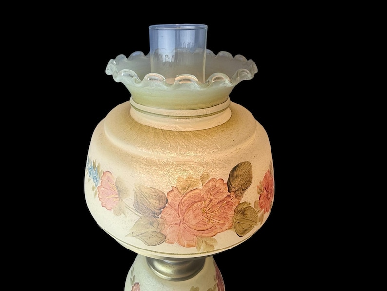 Puede incluir: Una l&aacute;mpara de aceite de estilo vintage con una base redondeada de color crema adornada con dise&ntilde;os florales rosas y melocot&oacute;n y hojas verdes. La l&aacute;mpara presenta una pantalla de vidrio verde claro con volantes y una chimenea de vidrio transparente. La l&aacute;mpara est&aacute; sobre un fondo negro.