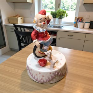 May include: A vintage ceramic clown figurine riding a unicycle. The clown is dressed in a red jacket, blue pants, and a white collar. A small white dog and a red box are also on the base. The base is white with pink polka dots and a pink scalloped edge.