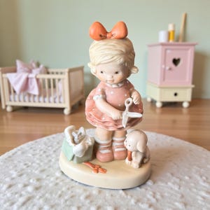 May include: A ceramic figurine of a young girl with a large orange bow in her hair, wearing a pink polka-dot dress, holding scissors. A small dog and a toy are at her feet. The figurine is on a cream-colored base.