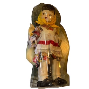 May include: A vintage doll in a clear plastic case. The doll wears a white dress with a red stripe, a floral patterned top, and a yellow scarf. The doll has black shoes and is holding a yellow sack.
