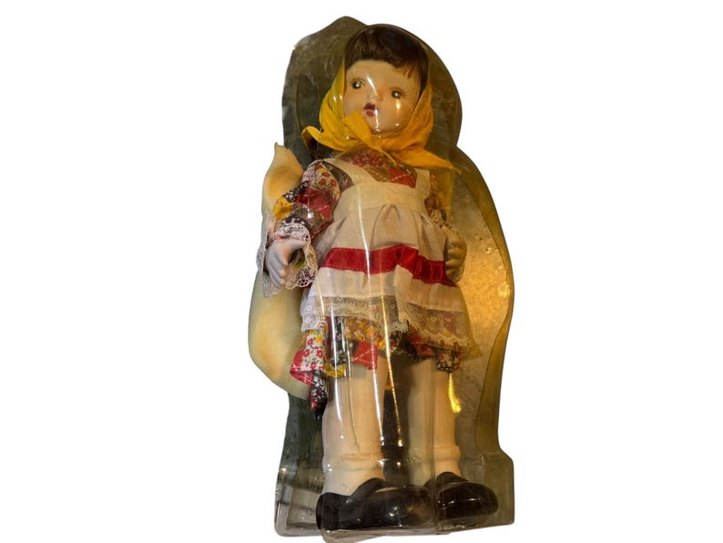 May include: A vintage doll in a clear plastic case. The doll wears a white dress with a red stripe, a floral patterned top, and a yellow scarf. The doll has black shoes and is holding a yellow sack.