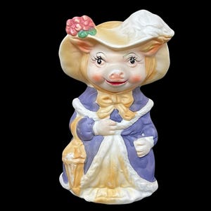 May include: A ceramic figurine of a pig wearing a yellow hat with a white trim and a purple dress. The pig has a pink face with a small pink nose and a pink bow around its neck.