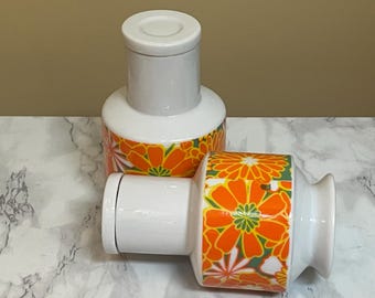 May include: Two white ceramic jars with orange, green, and white floral designs. The jars have lids and are perfect for storing small items.