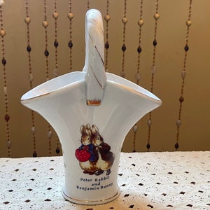 May include: White ceramic vase with a handle shaped like a basket. The vase features a depiction of Peter Rabbit and Benjamin Bunny with a red strawberry. The text "Peter Rabbit and Benjamin Bunny" is printed on the vase.