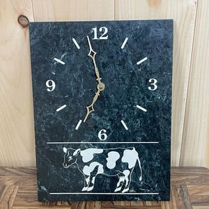 May include: A green marble clock with white numbers and a cow design. The clock face reads 12, 3, 6, and 9. The cow is white with black spots.