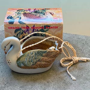 May include: A white ceramic swan hanging planter with green and brown wing details, featuring a small floral accent on the chest and a natural fiber rope for suspension. The planter is displayed in front of its original cardboard box packaging.