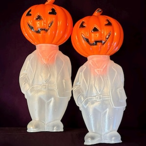 May include: Two molded plastic figures featuring bright orange pumpkin heads with black painted facial features. Each figure has a translucent white body shaped like a person wearing a suit, tie, and jacket, standing upright with hands in pockets.