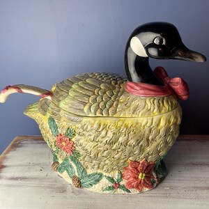 May include: A ceramic goose-shaped serving bowl with a red ribbon around its neck. The bowl is decorated with green and brown feathers, and has a detailed floral design with red and yellow flowers. The bowl has a removable lid with a handle shaped like a candy cane.