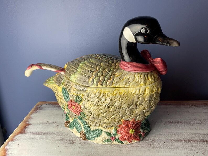 May include: A ceramic goose-shaped serving bowl with a red ribbon around its neck. The bowl is decorated with green and brown feathers, and has a detailed floral design with red and yellow flowers. The bowl has a removable lid with a handle shaped like a candy cane.