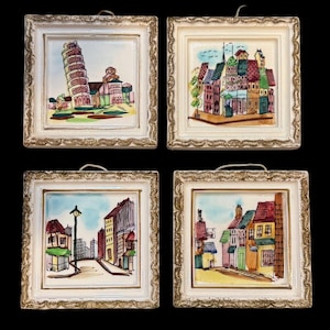 May include: Four framed ceramic tiles with hand-painted scenes of European cityscapes. The artwork includes the Leaning Tower of Pisa, colorful buildings, and street scenes. Each tile is set in a white frame with a decorative gold trim.