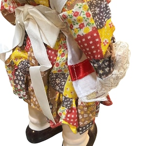 May include: A vintage doll wearing a patchwork dress with red, yellow, and brown floral patterns. The doll has white lace trim and a white ribbon tied at the waist. The doll's shoes are black.