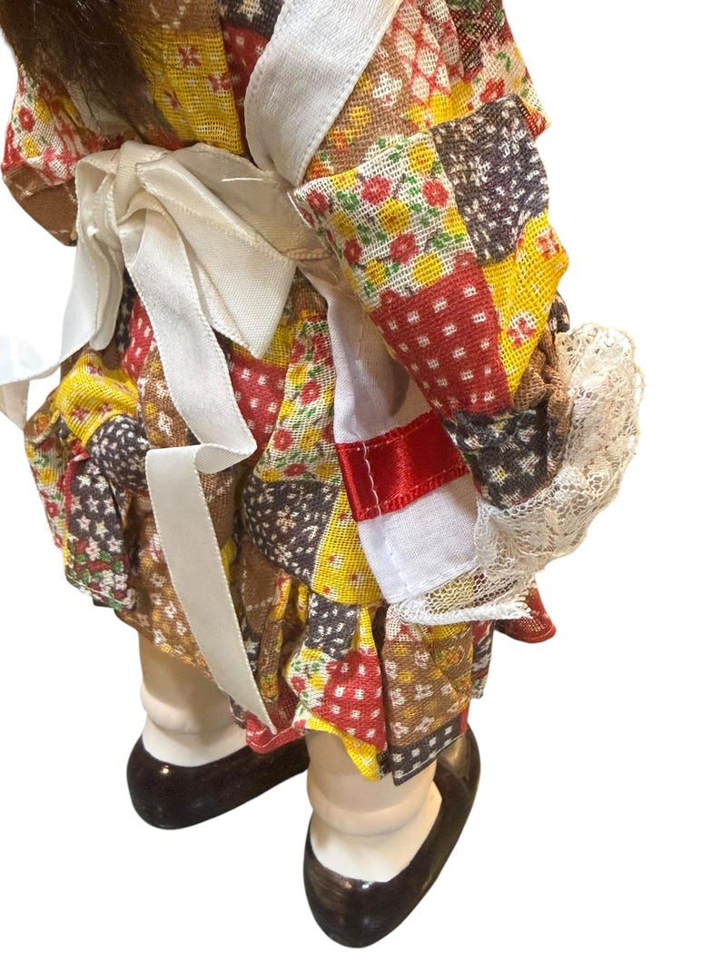 May include: A vintage doll wearing a patchwork dress with red, yellow, and brown floral patterns. The doll has white lace trim and a white ribbon tied at the waist. The doll's shoes are black.