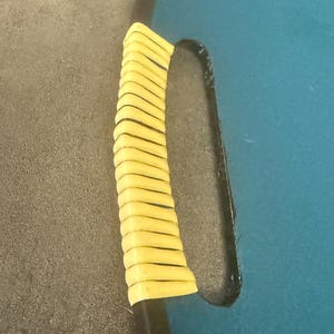 May include: Close-up of a black handle with a yellow grip. The handle is attached to a teal blue surface. The grip is made of multiple yellow, horizontal, rectangular pieces.