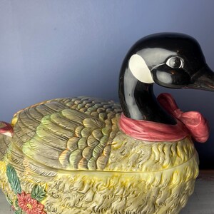 May include: A ceramic goose-shaped serving dish with a red and white bow around its neck. The goose is decorated with brown, green, and yellow feathers and has a floral design on its side.