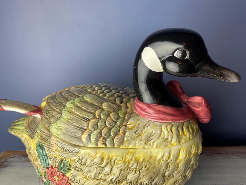 May include: A ceramic goose-shaped serving dish with a red and white bow around its neck. The goose is decorated with brown, green, and yellow feathers and has a floral design on its side.