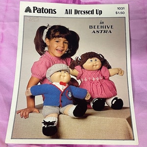 Puede incluir: Un folleto de patrones de tejido de punto con dos muñecas vestidas con atuendos tejidos. Las muñecas llevan atuendos rosas y azules con detalles blancos. La portada del folleto presenta el texto "Patons All Dressed Up in BEEHIVE ASTRA 1031 $1.50".