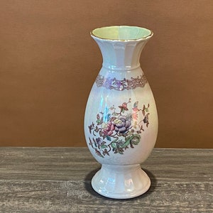 May include: A white ceramic vase with a pearlescent finish and a floral design. The vase has a wide mouth and a round base.