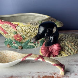 May include: A ceramic goose-shaped serving bowl with a green, brown, and yellow feathered design. The goose is wearing a red bow and the bowl has a matching ladle.