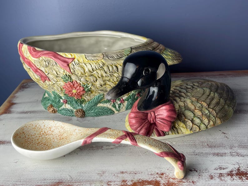 May include: A ceramic goose-shaped serving bowl with a green, brown, and yellow feathered design. The goose is wearing a red bow and the bowl has a matching ladle.
