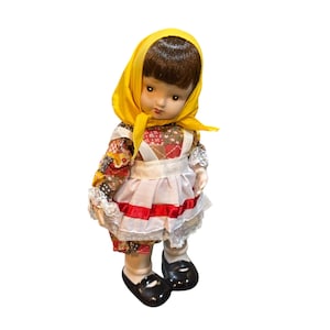 May include: A vintage doll with brown hair and a yellow headscarf. The doll wears a colorful patchwork dress, a white apron with red trim, and black shoes. The doll's face has a slightly sad expression.