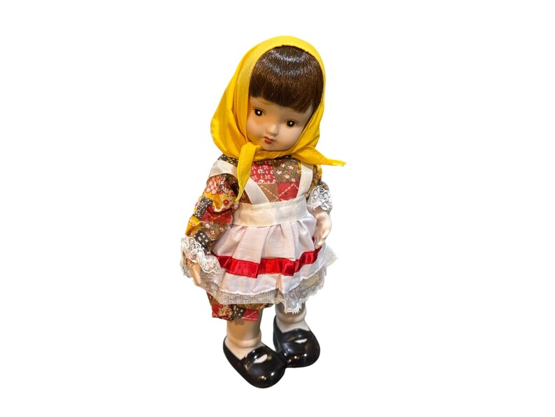 May include: A vintage doll with brown hair and a yellow headscarf. The doll wears a colorful patchwork dress, a white apron with red trim, and black shoes. The doll's face has a slightly sad expression.